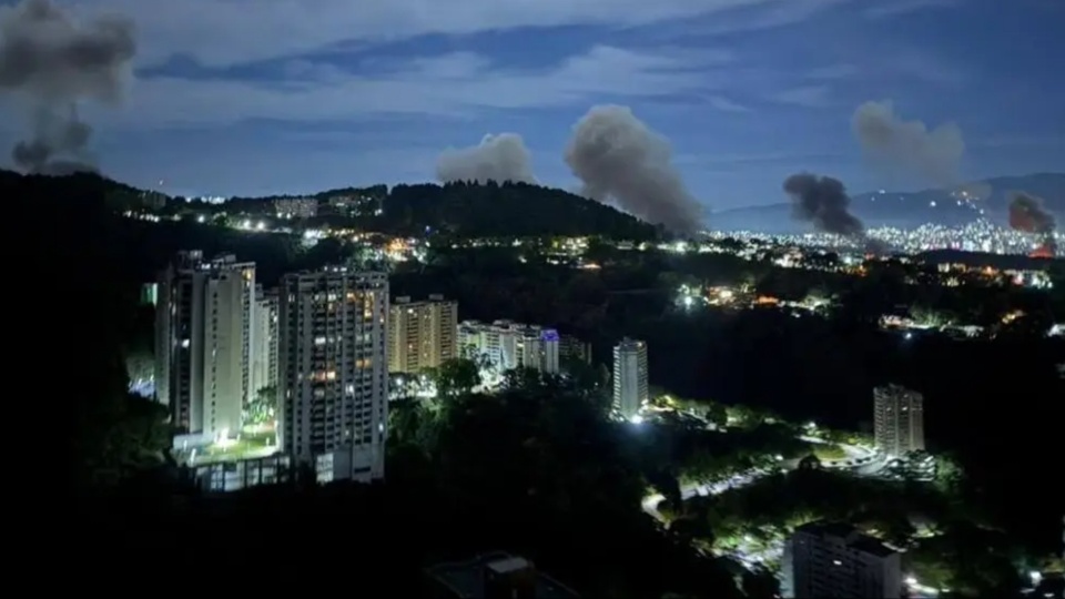 A picture of the city of Caracas, Venezuela, at night. Plumes of smoke rise into the air from US bombing.